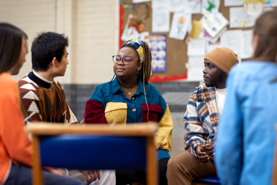 A group of young people talking to each other. They are sitting in a circle and look like they are in a school setting.
