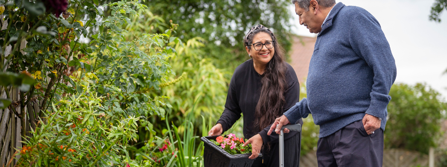 An older man using a walking cane and a younger woman gardening together.