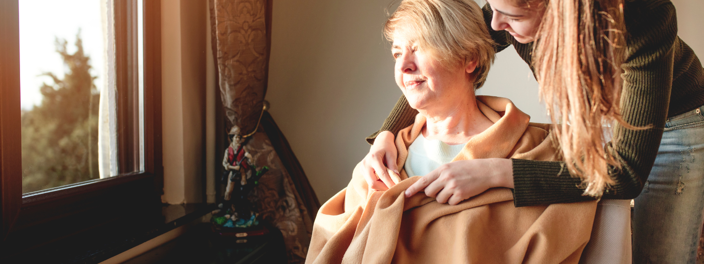 A younger woman wrapping a blanket around an older woman. The older woman is sitting in a chair and looking out of a window.