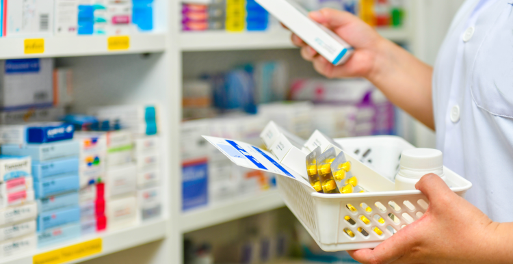 Close up showing a pharmacist's hands and arms. They are filling a prescription basket, standing in front of shelves containing medication boxes.