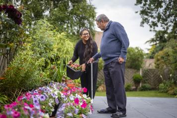 An older man using a walking stick and a younger woman gardening.