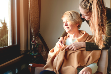 A younger woman wrapping a blanket around an older woman. The older woman is sitting in a chair and looking out of a window.