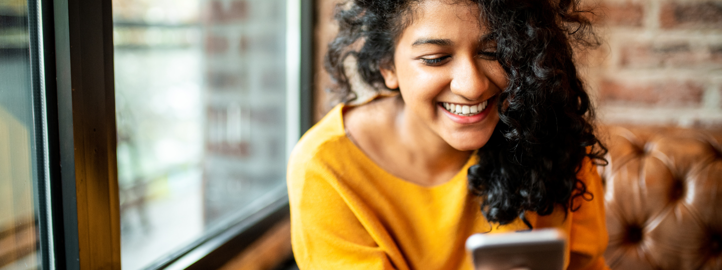 A woman sitting in a cafe, looking at a mobile phone and smiling.