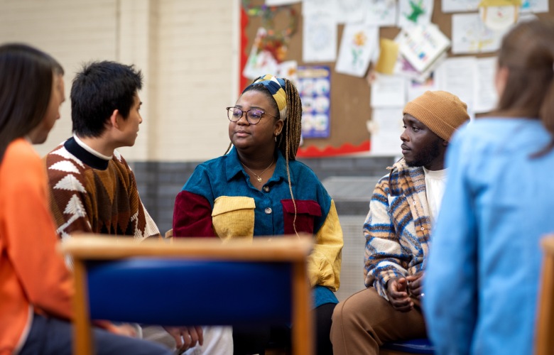 A group of young people talking to each other. They are sitting in a circle and look like they are in a school setting.