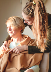 A younger woman wrapping a blanket around an older woman. The older woman is sitting in a chair and looking out of a window.